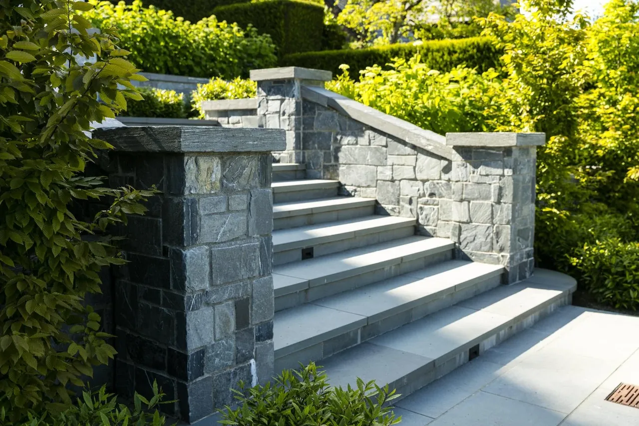 Stone stairway with solid stone walls in a landscaped outdoor setting with dense greenery.