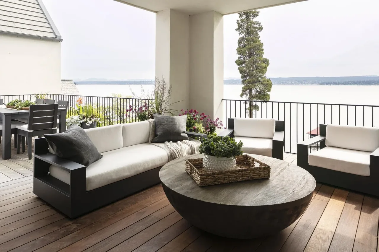 Covered outdoor living space featuring cushioned black-framed sofas and chairs with a round wooden coffee table and a lake view.