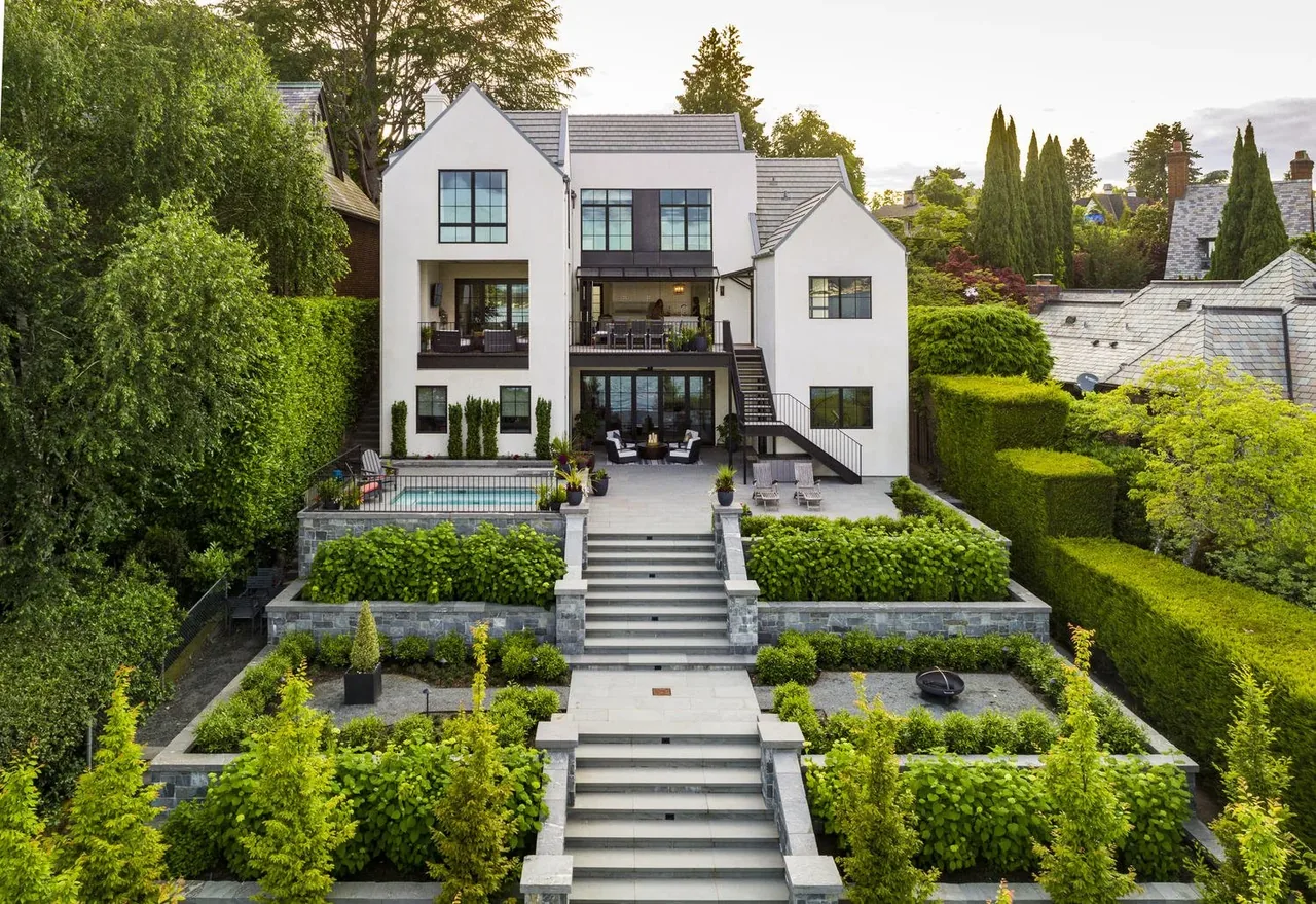 Rear exterior of Laurelhurst Contemporary house by Paul Moon Design featuring white facade, black window frames, multi-level patio, and formal garden.