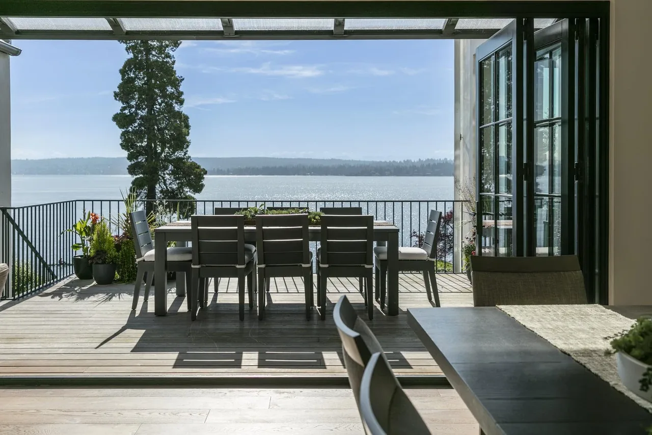 Outdoor dining area with modern furniture on a wood deck overlooking water, designed by Paul Moon Design.