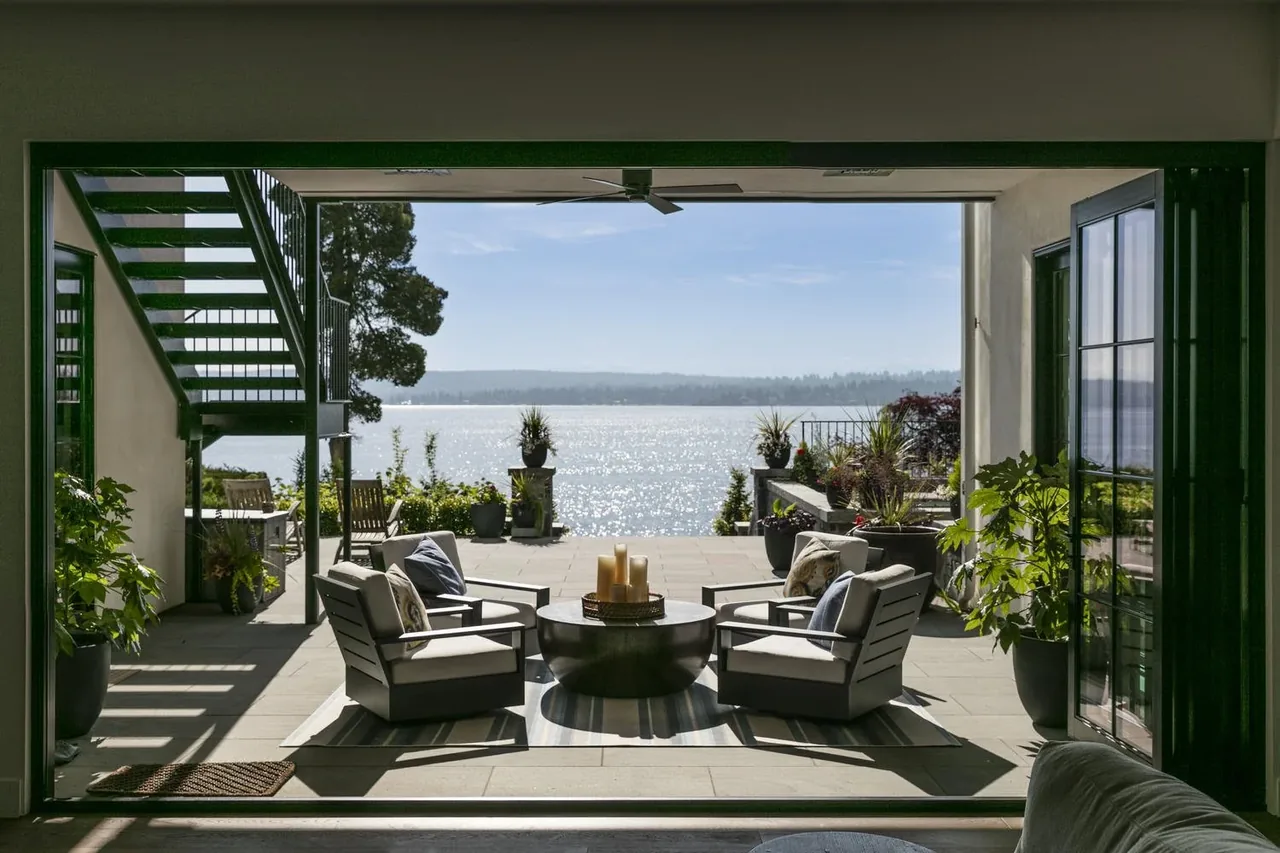 Outdoor patio with seating area faces a lake view, featuring light stone flooring and potted plants.