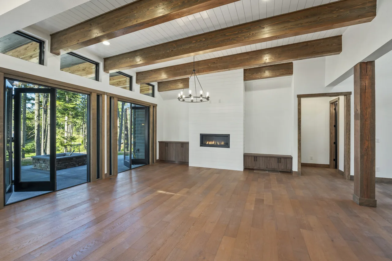 Room features exposed wooden beams, floor-to-ceiling glass doors, and a linear fireplace with built-in cabinetry.