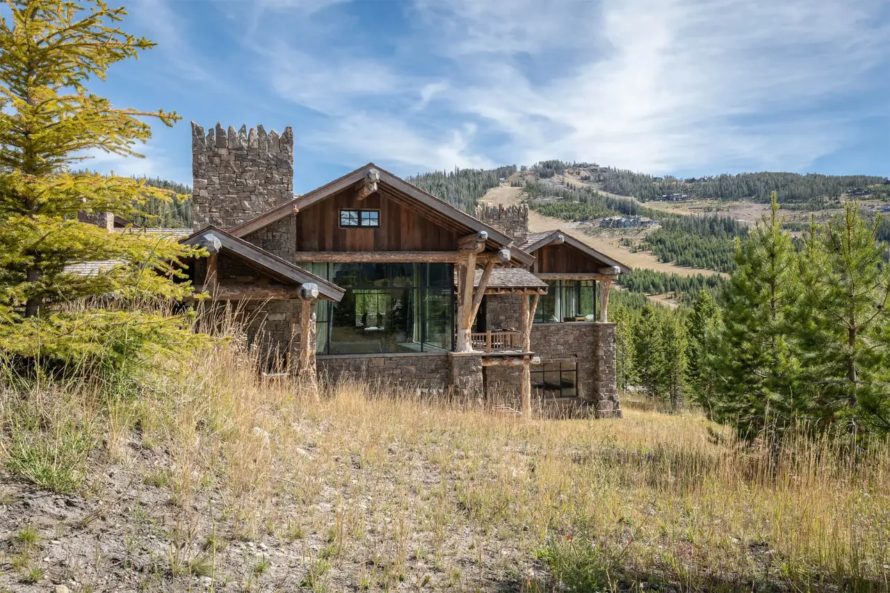 Chalet Redux features a stone and wood exterior with large glass windows and a mountain backdrop.