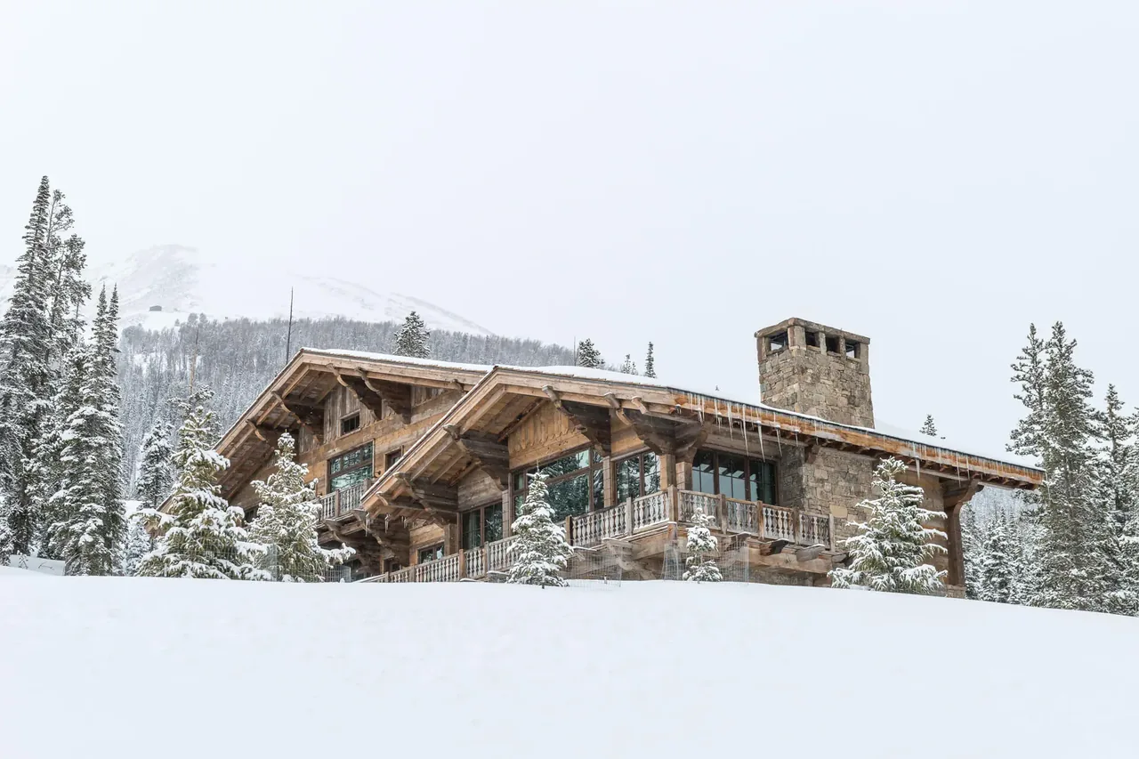 Winter exterior view of Chalet Redux, a single-story rustic chalet with large stone chimney and wood beams.