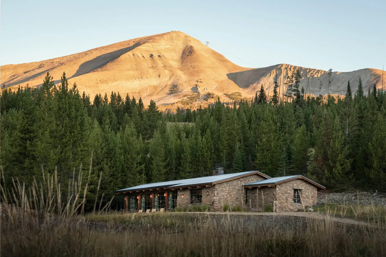 Chalet Redux by Pearson Design Group is a stone house with metal gable roofs nestled near forest and mountain.