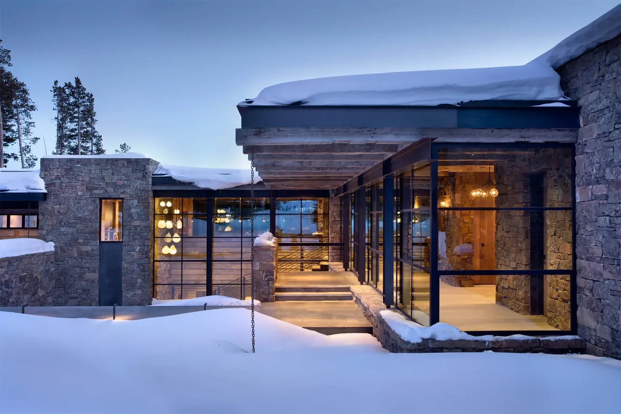 Chalet Redux features stone walls paired with large black-framed glass windows under a flat roof covered with snow.