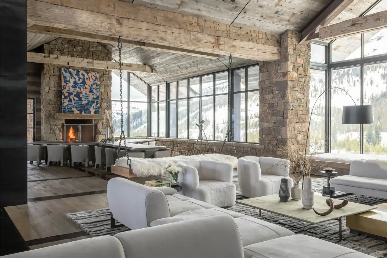 Chalet Redux living room features stone fireplace, floor-to-ceiling windows with mountain views, and white upholstered seating.