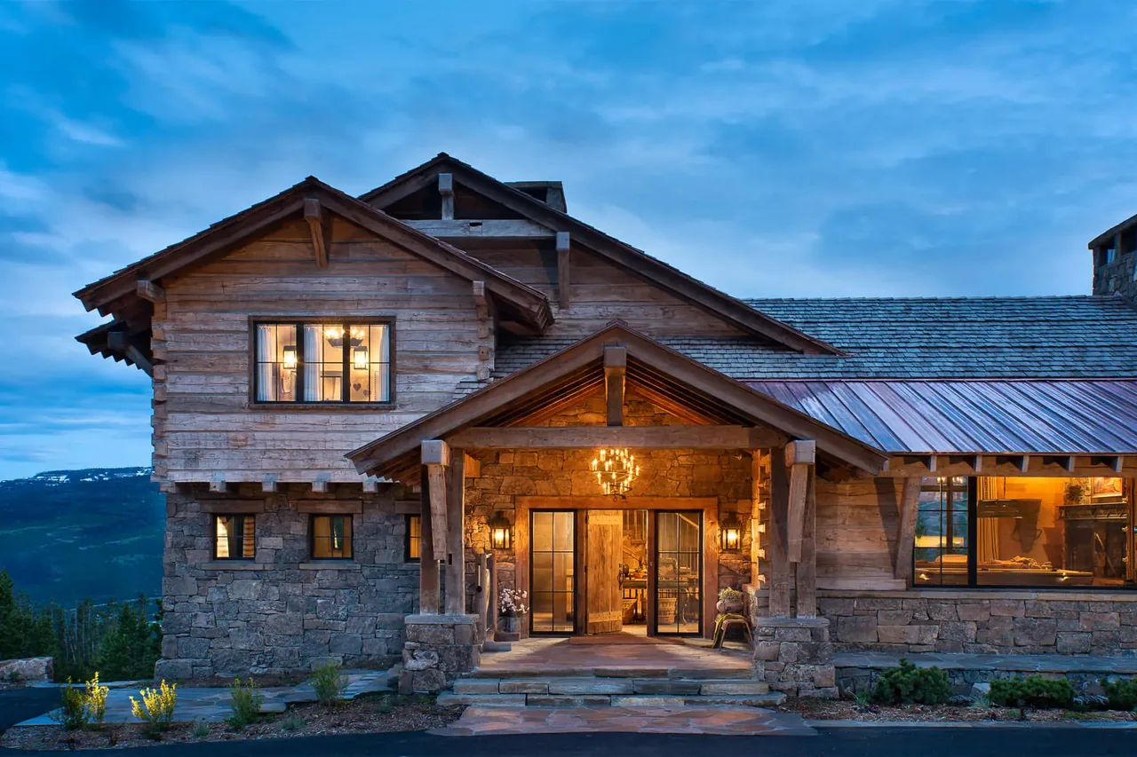 Single-story chalet featuring stone pillars, a wood exterior, and warm interior lighting visible through large windows at dusk.