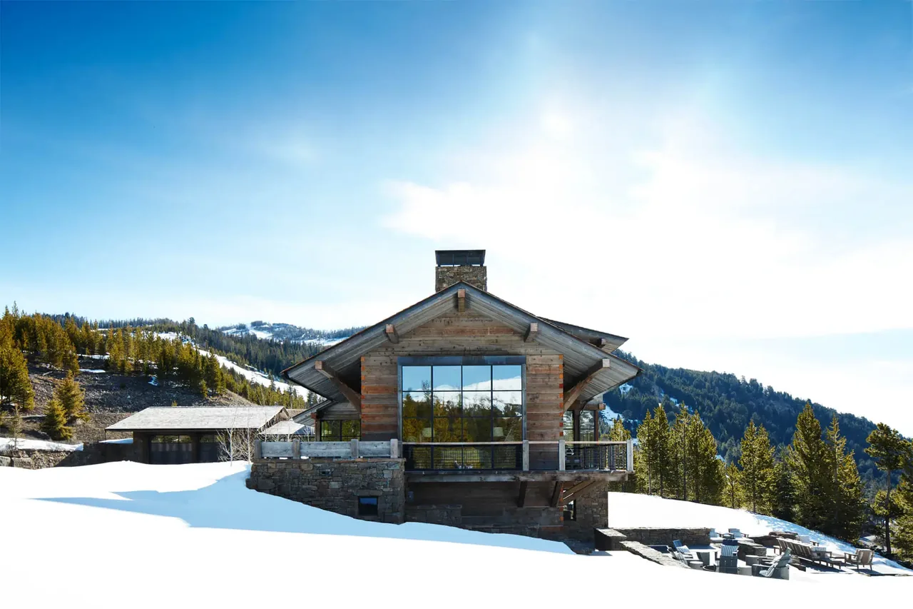 Chalet Redux by Pearson Design Group features wood and stone construction with a prominent central window facing a snow-covered outdoor space.