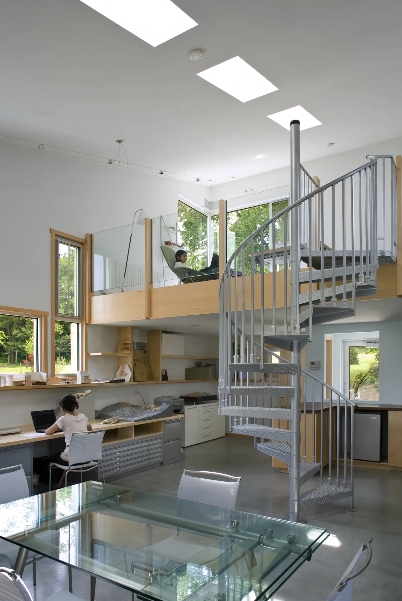 Modern home office features a metal spiral staircase, glass railing on upper loft, and large windows providing natural light.