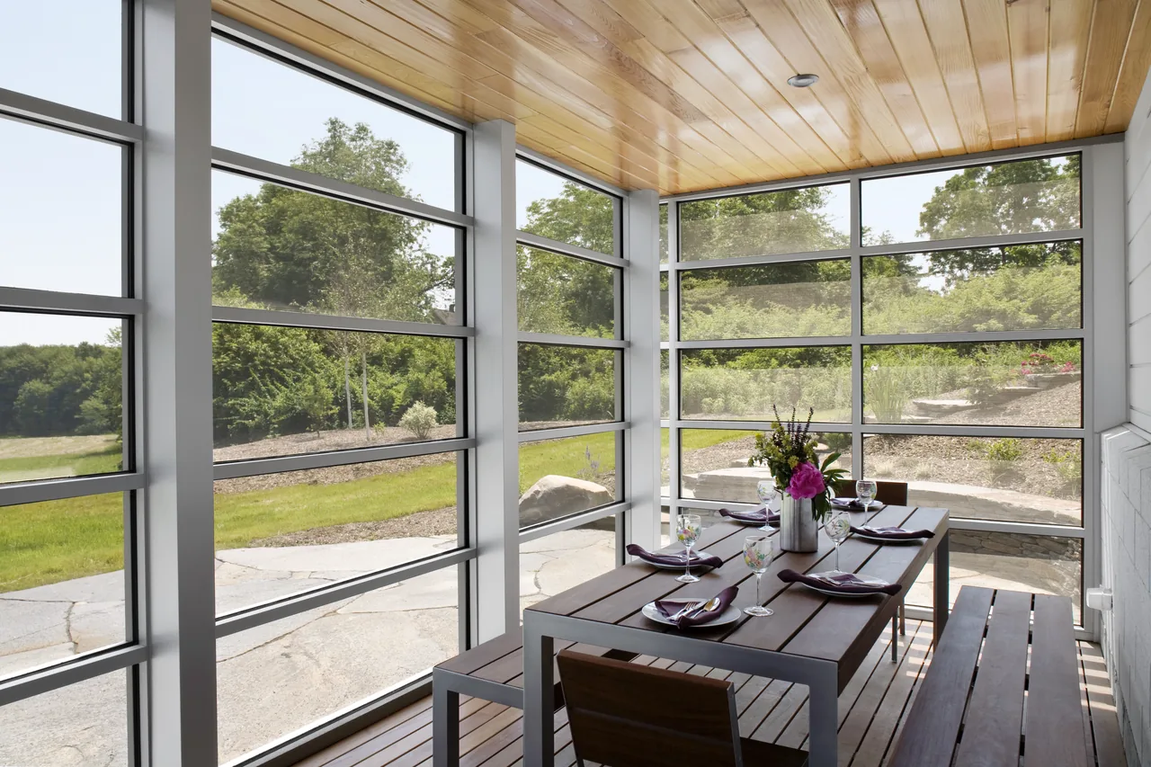 Dining space in a screened porch with metal-framed windows overlooking a landscaped garden area.