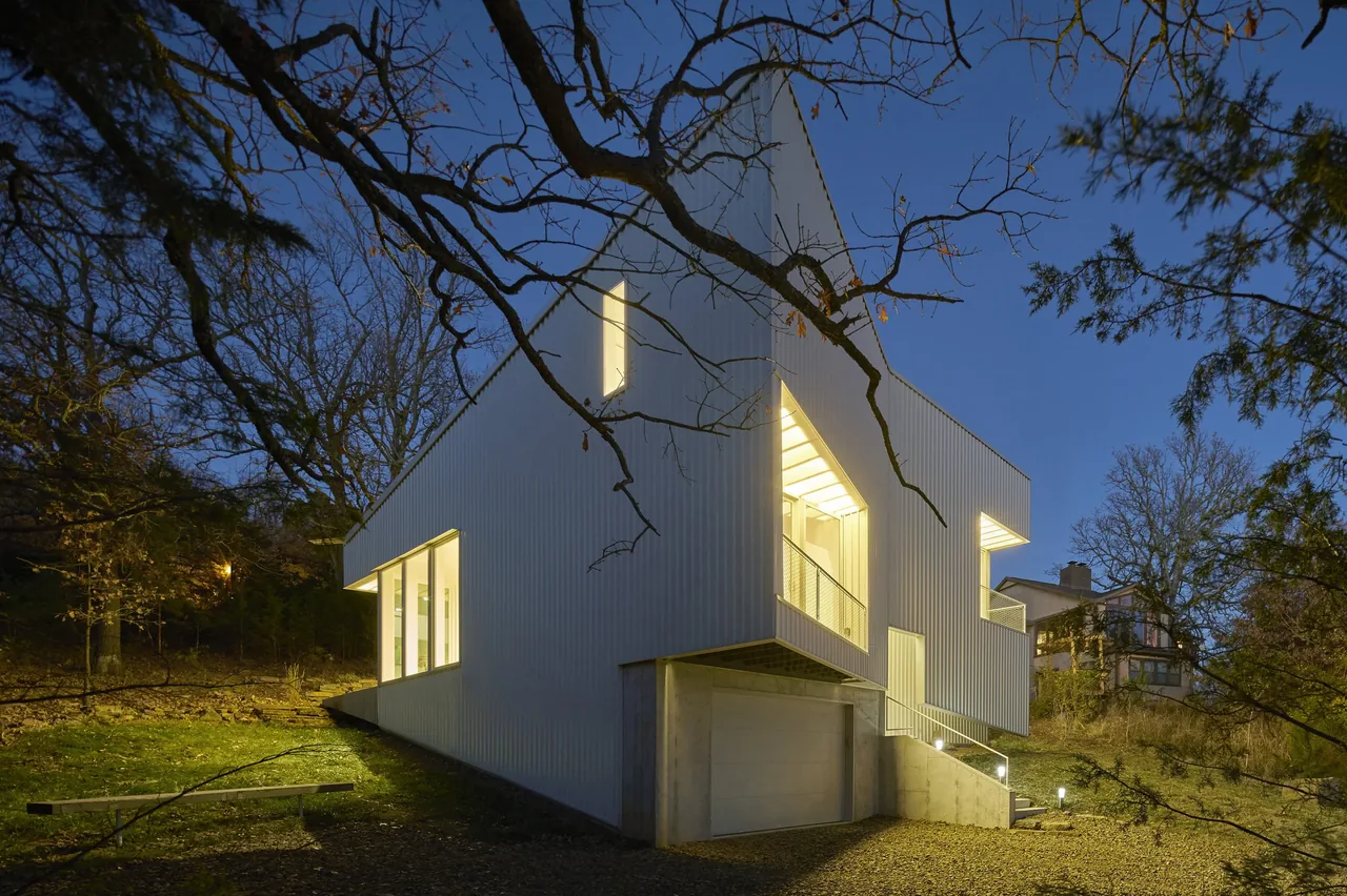 Modern hillside house with white vertical siding and large illuminated windows at night.
