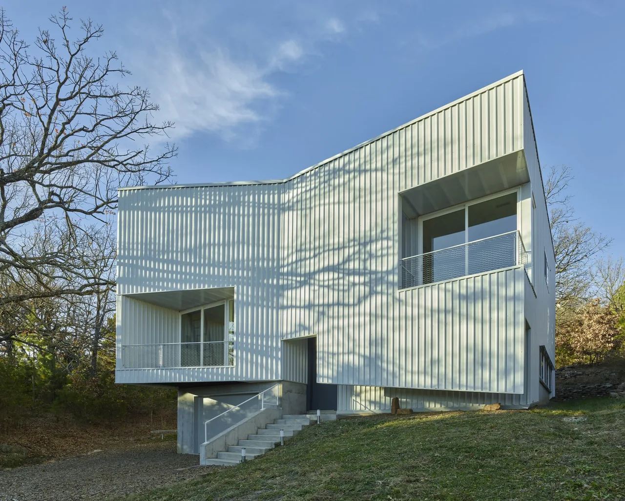 The Hillside Rock project features a modern house with white metal siding and cantilevered sections, situated on a wooded hillside.