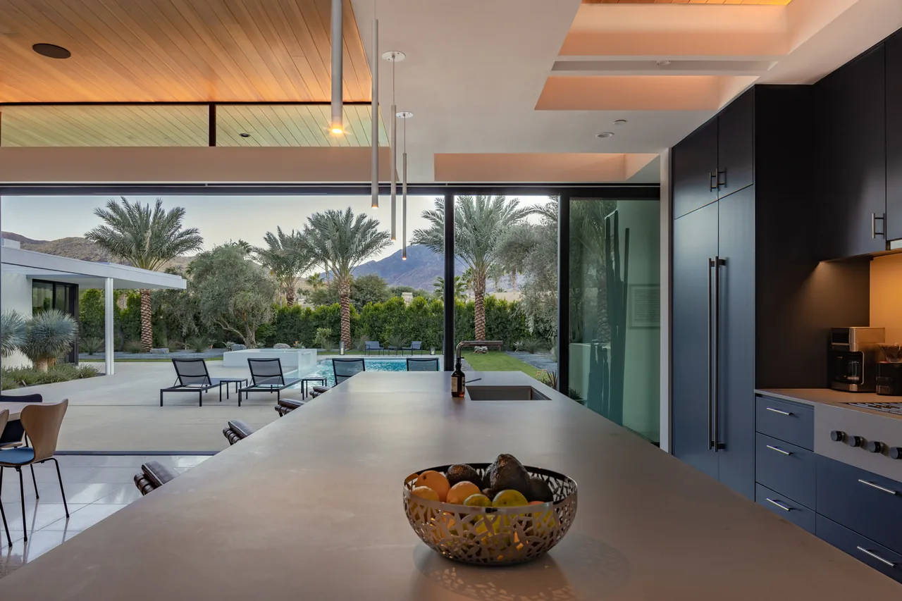 Kitchen with a large island and glass wall opens to a backyard with pool and palm trees, mountains visible in the distance.