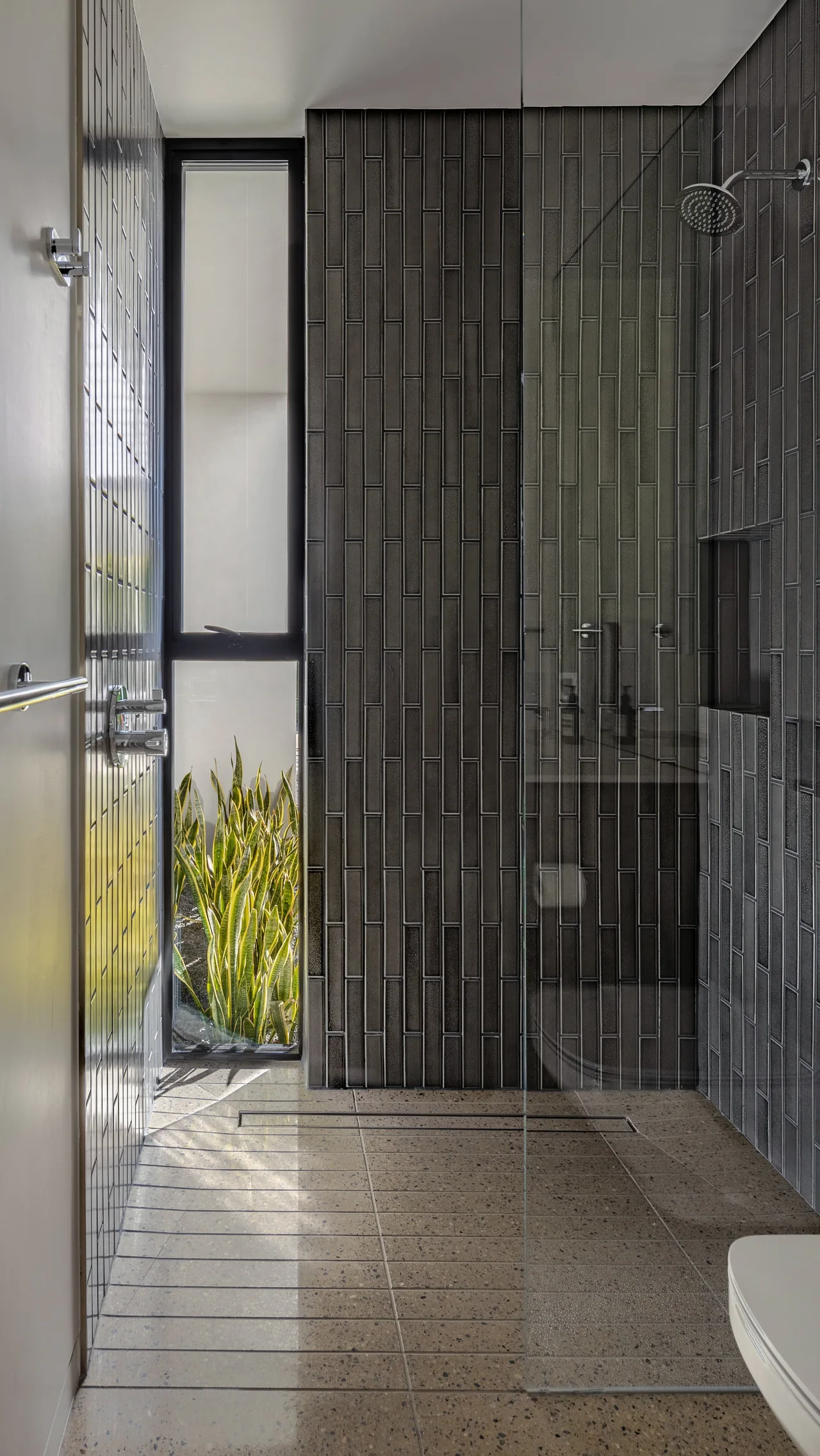 The Sky Ridge shower area features black vertical tiles, a rain showerhead, and a clear glass panel next to a tall window with greenery outside.