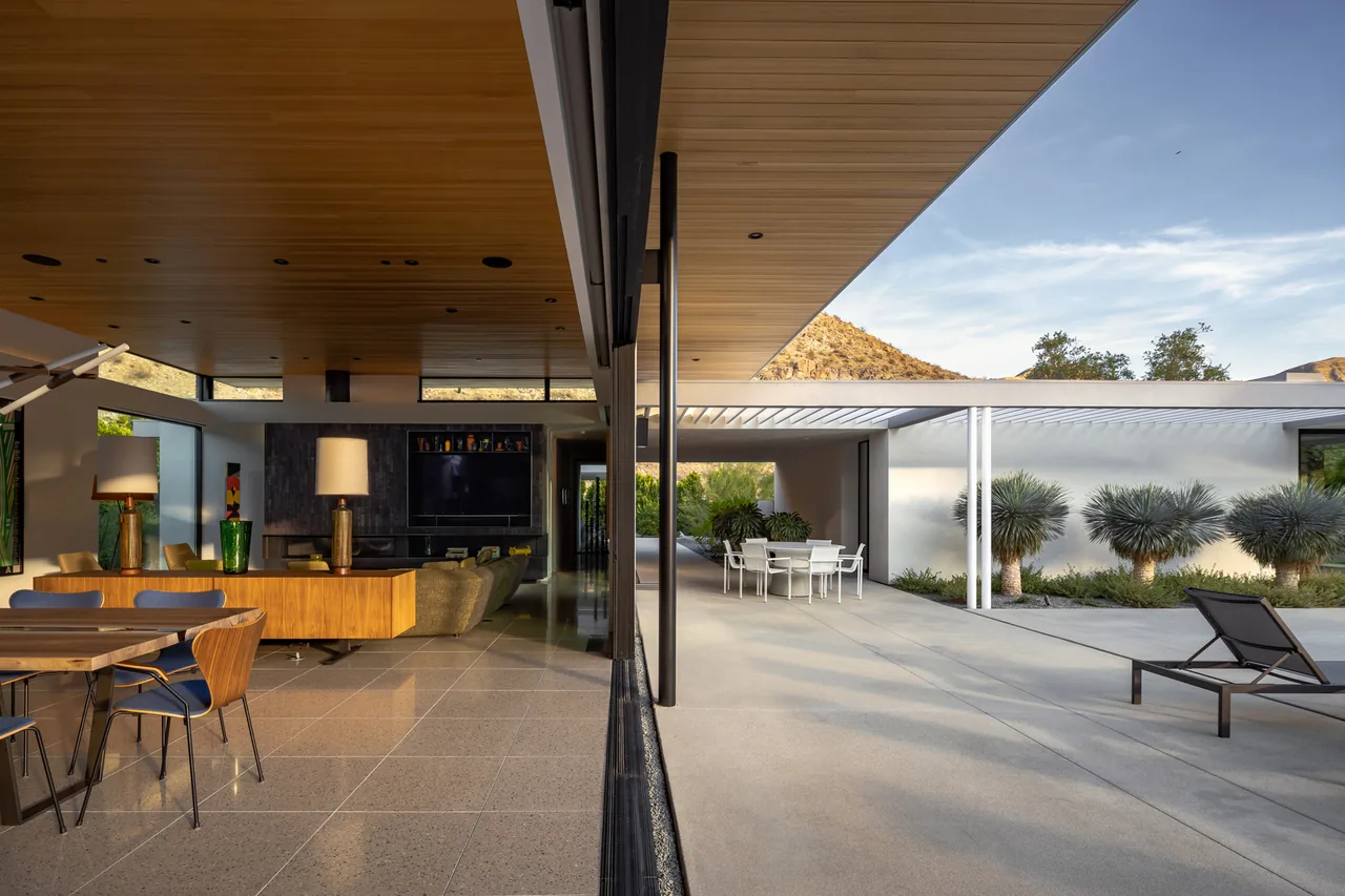 Modern home interior with wood ceiling and polished floor opening to a patio with outdoor seating under a pergola and desert landscaping.