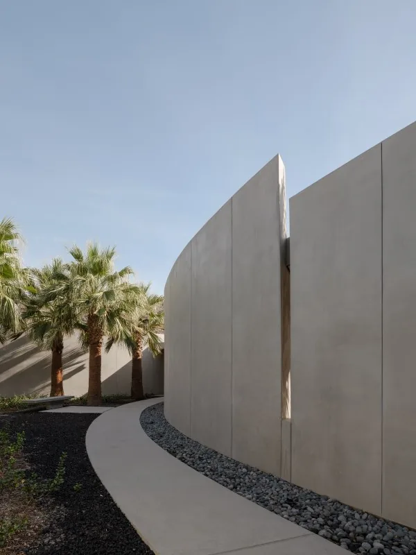 A curved concrete wall with a thin vertical window creates a path alongside desert landscaping with palm trees at Eldorado Tres.