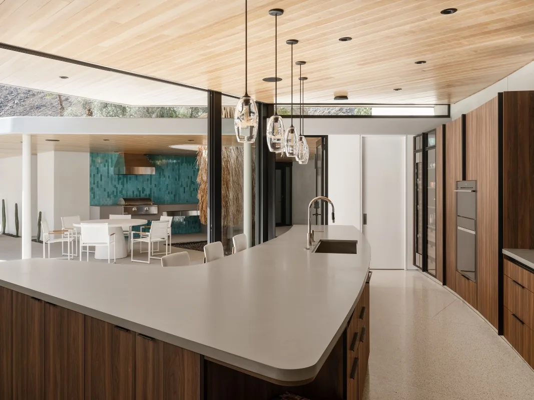 Kitchen features a curved island with pendant lights and overlooks an outdoor dining area with a turquoise tiled backsplash.