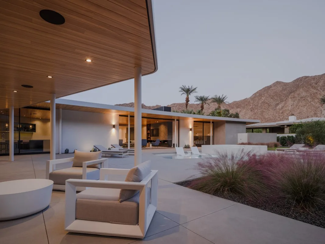 Modern outdoor living space with curved wood ceiling, pool, and desert landscaping next to a flat-roofed house.