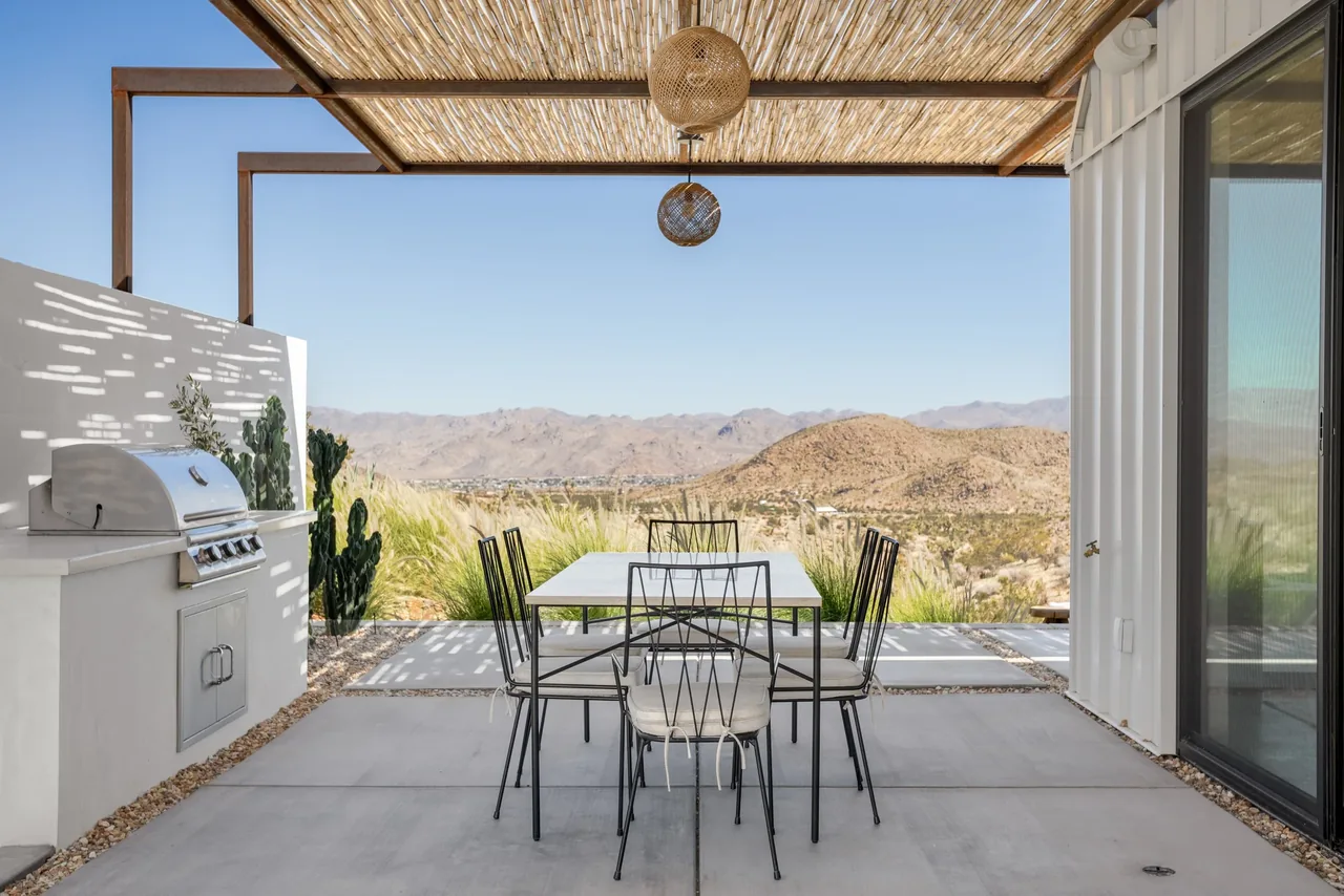 Outdoor dining space at Alta guesthouse with metal furniture and pergola roof, overlooking a desert landscape with mountains.