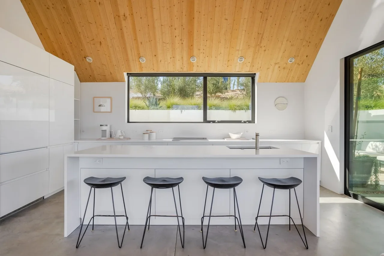 Minimalist white kitchen features a vaulted wood ceiling and large black-framed window overlooking greenery.