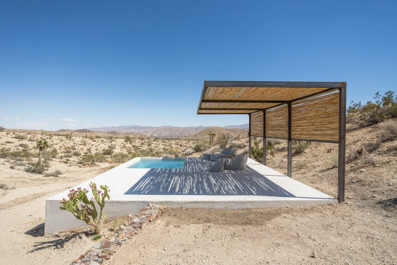 Alta Guesthouse features a minimalist concrete patio with a pergola and pool against a desert setting.