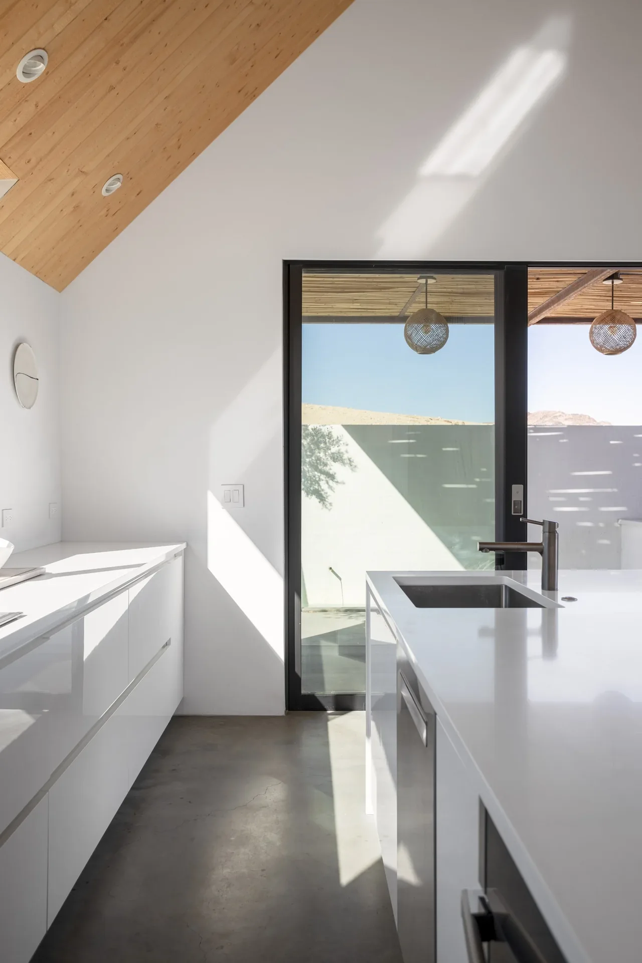 Kitchen of alta guesthouse features white cabinetry, polished concrete floor, wood ceiling, and black-framed sliding glass door to outdoor space.