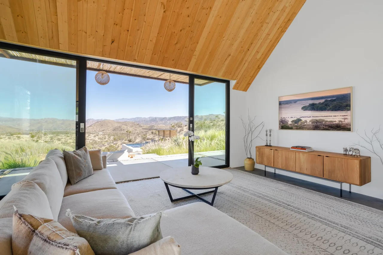 Living room with floor-to-ceiling glass doors opening to a scenic mountain view, featuring a wood-paneled vaulted ceiling and a minimalist wooden console.