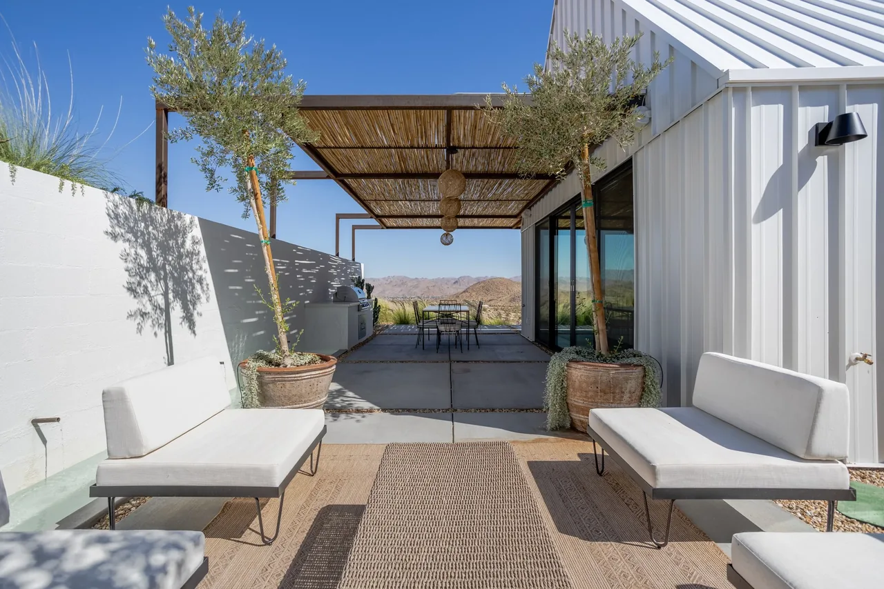 Patio with reed-shaded pergola, seating, and white metal siding on the guesthouse exterior.