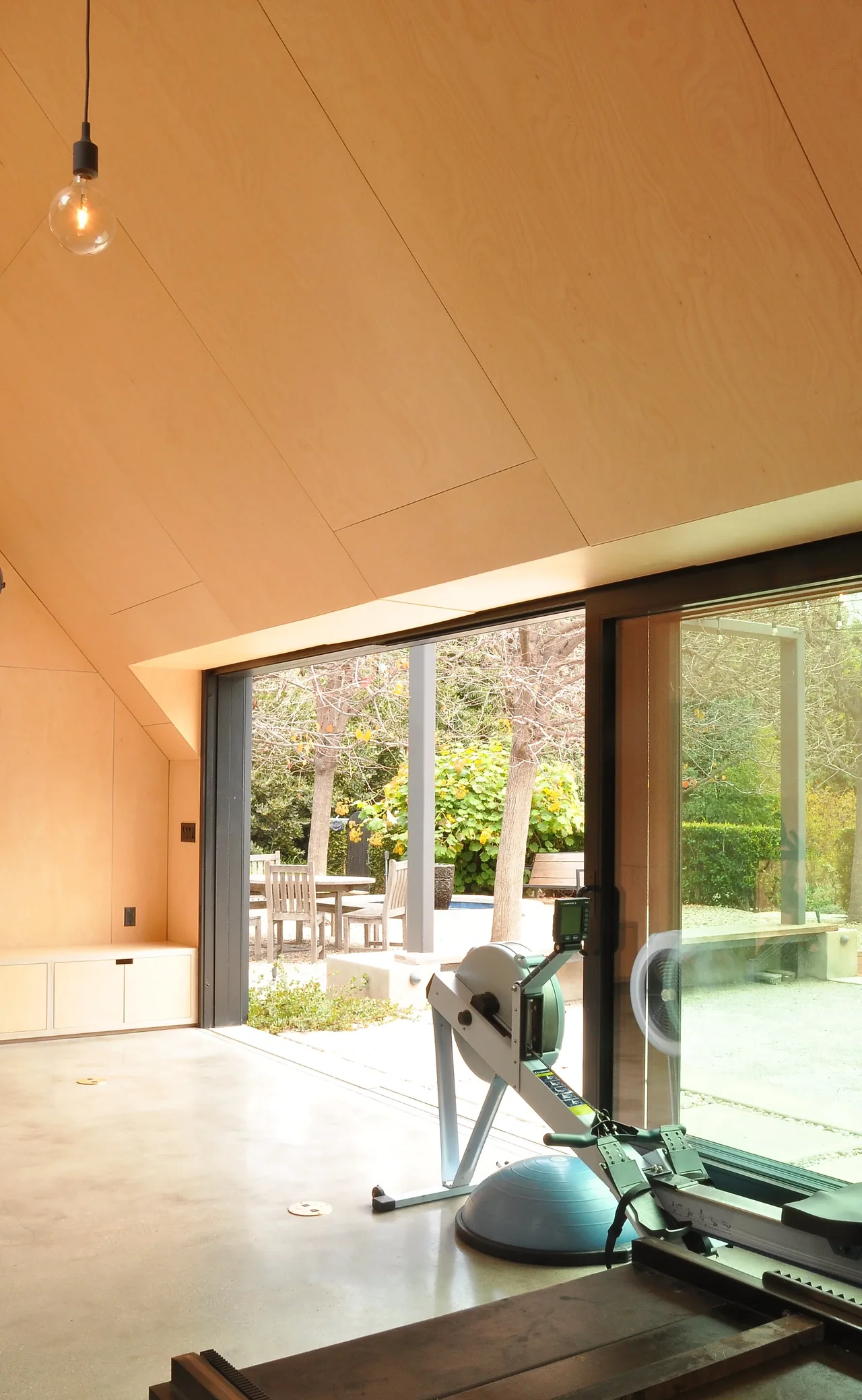 Exercise room with plywood panel ceiling and floor-to-ceiling sliding glass doors overlooking an outdoor seating area.