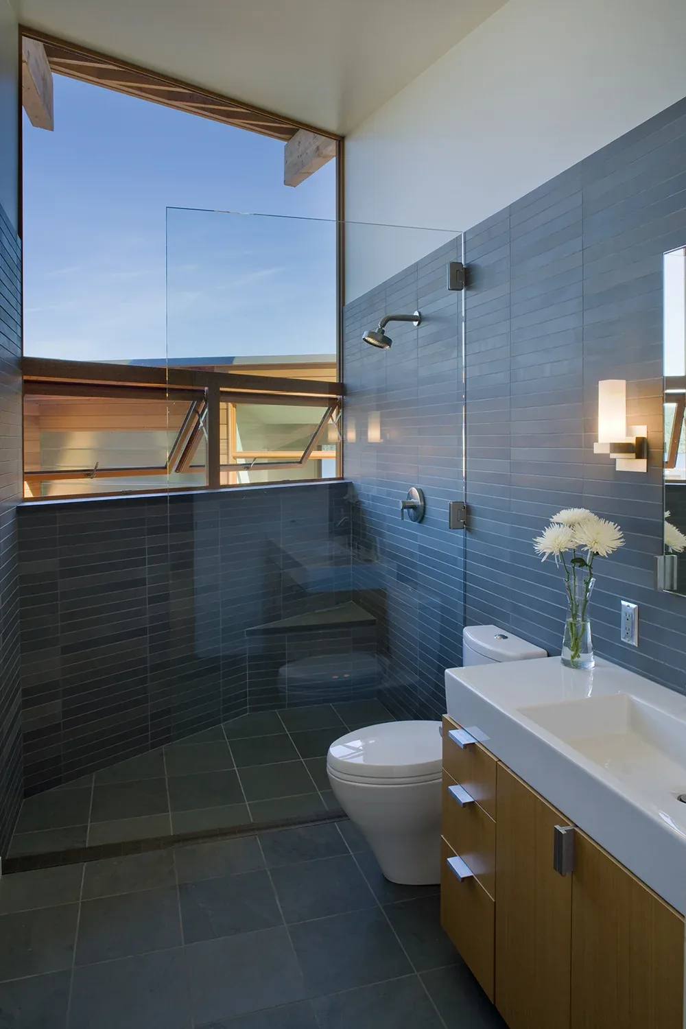 Modern bathroom with gray tiled walk-in shower, toilet, and wooden vanity under a large skylight window.
