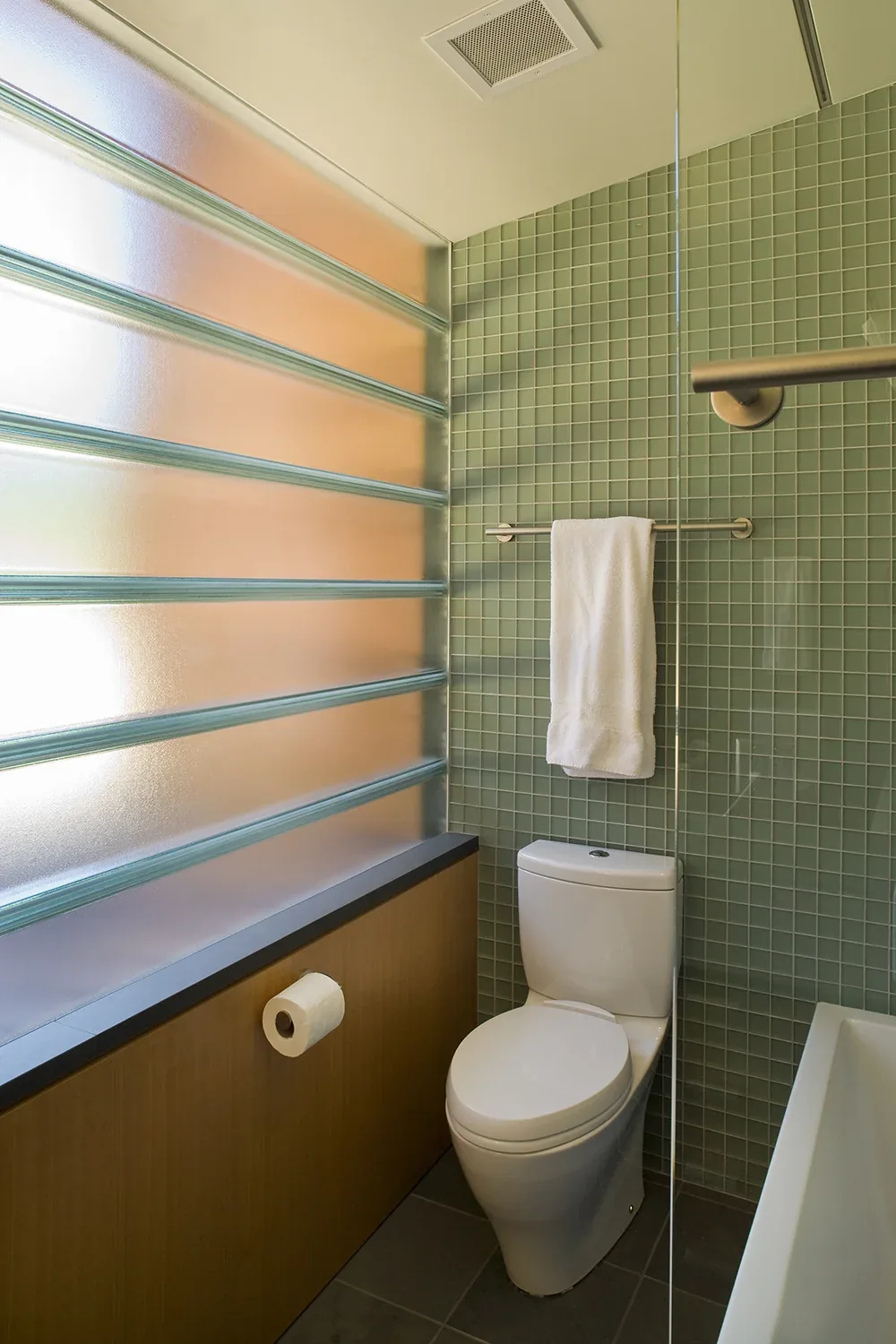 A bathroom in Y House features a toilet and a green tiled wall with a towel rack beside a frosted glass block window.