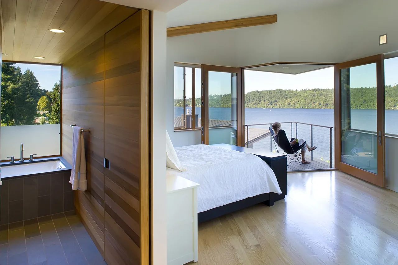 Master bedroom with hardwood floors opens to a balcony via folding glass doors, offering views of a lake, with bathroom and wood wall nearby.