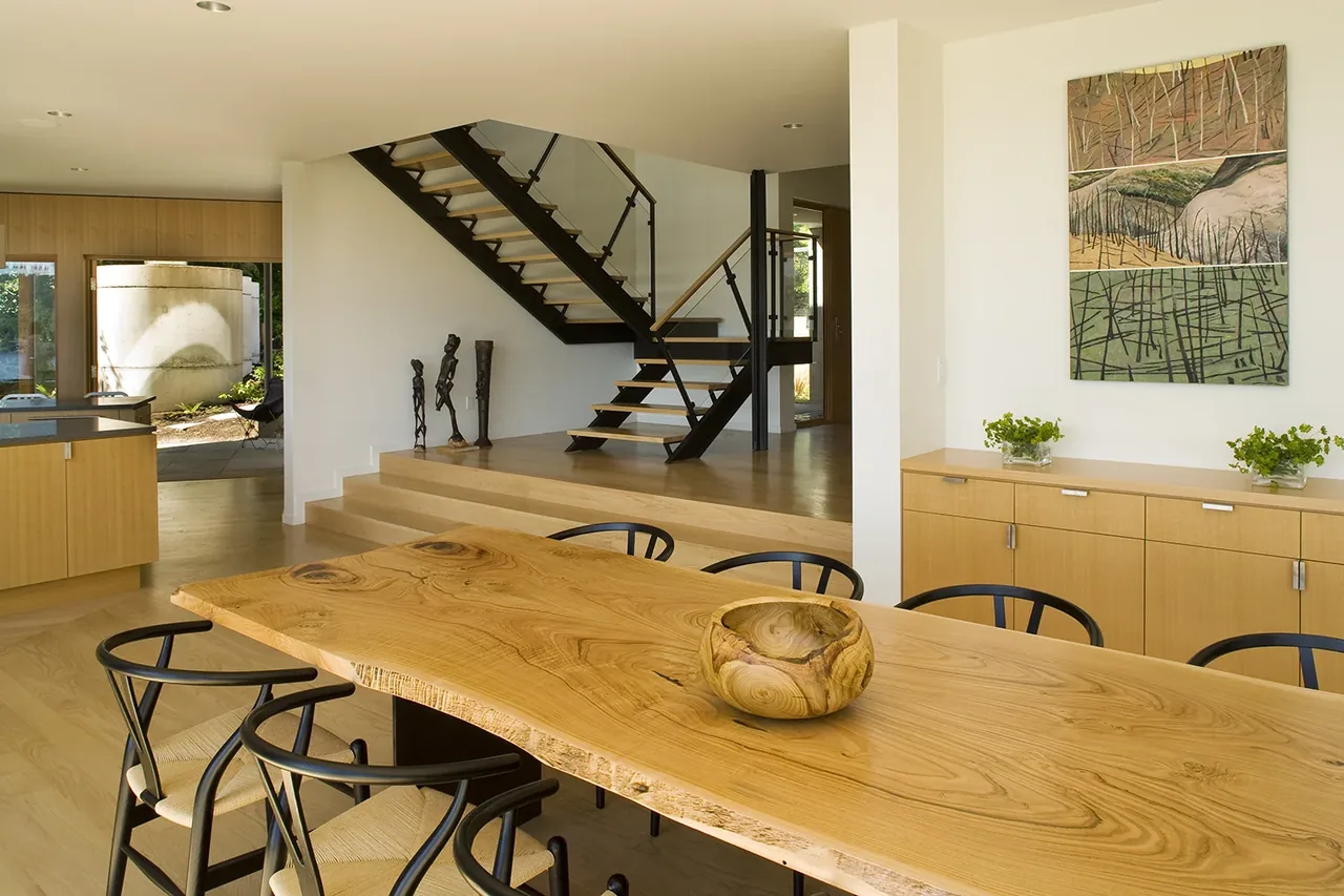 The dining area features a live-edge wooden table and black chairs, adjacent to a floating staircase with a glass balustrade.