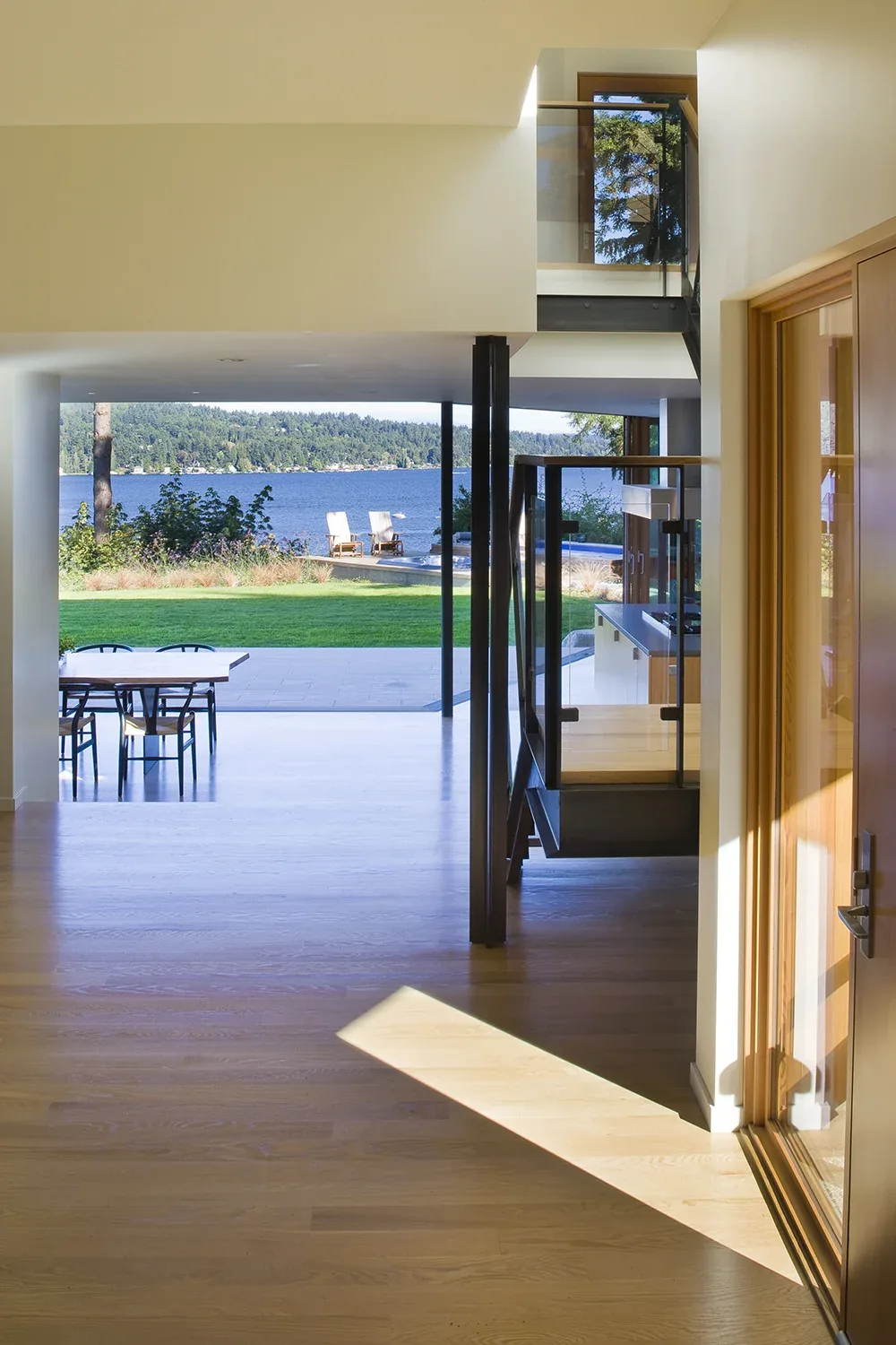 Interior view of Y House looking through to dining area and lake beyond with glass stair railing and wood flooring.