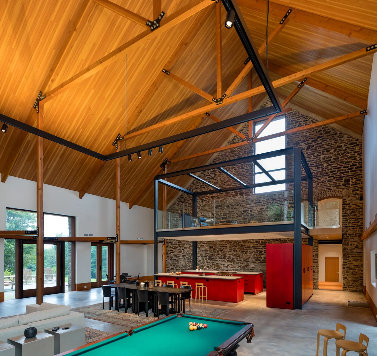 Barn-style interior features exposed wooden trusses, stone masonry wall, and an open kitchen with bold red cabinetry under a loft.
