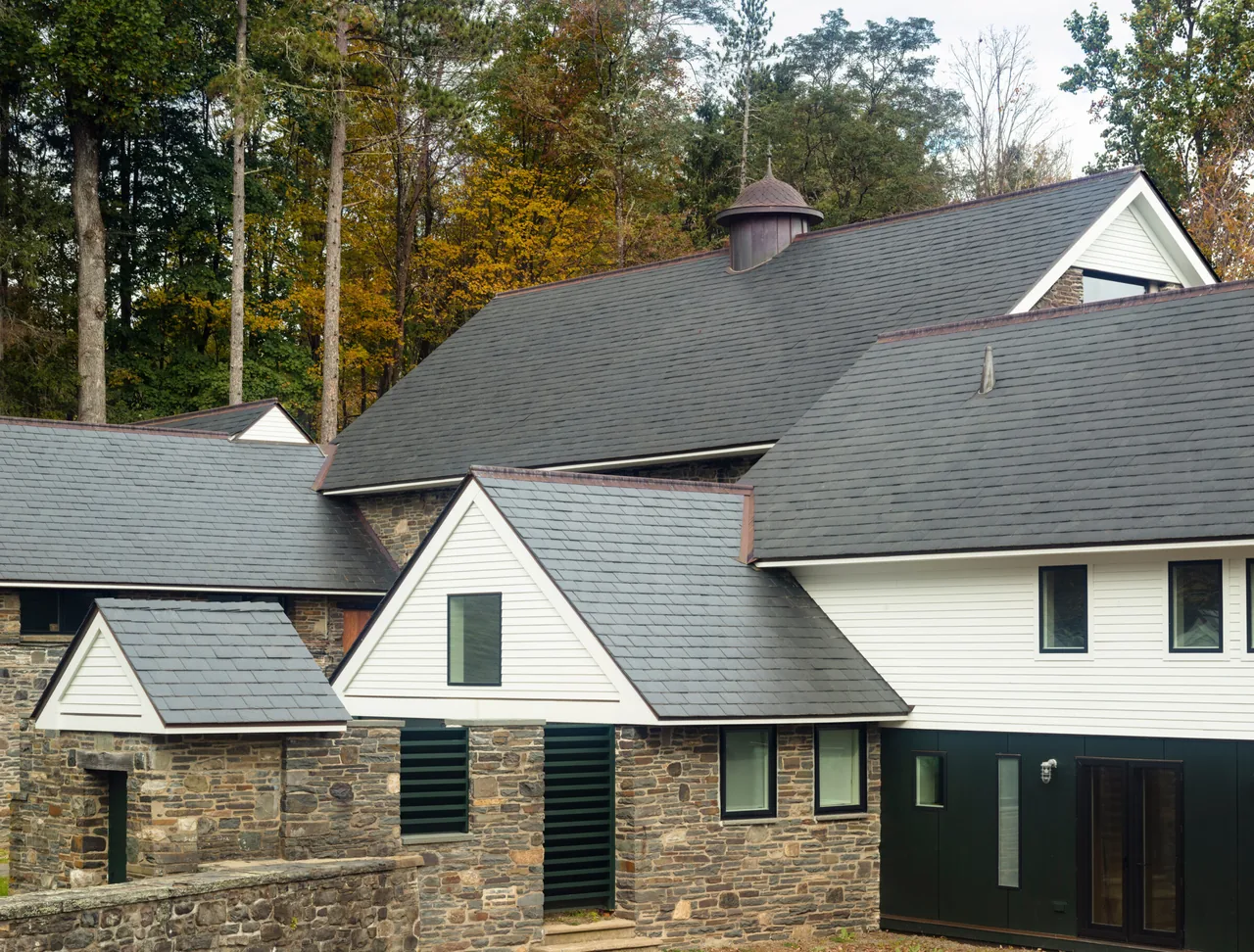 Barn with Aedicule features stone walls, slate roofs, and white siding with small modern windows, set among trees.