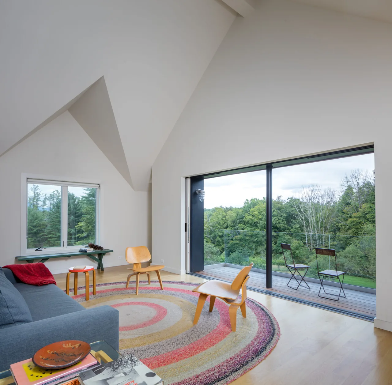 Living room with vaulted ceiling, light wood floor, and sliding glass door leading to a balcony with glass railing and outdoor chairs.