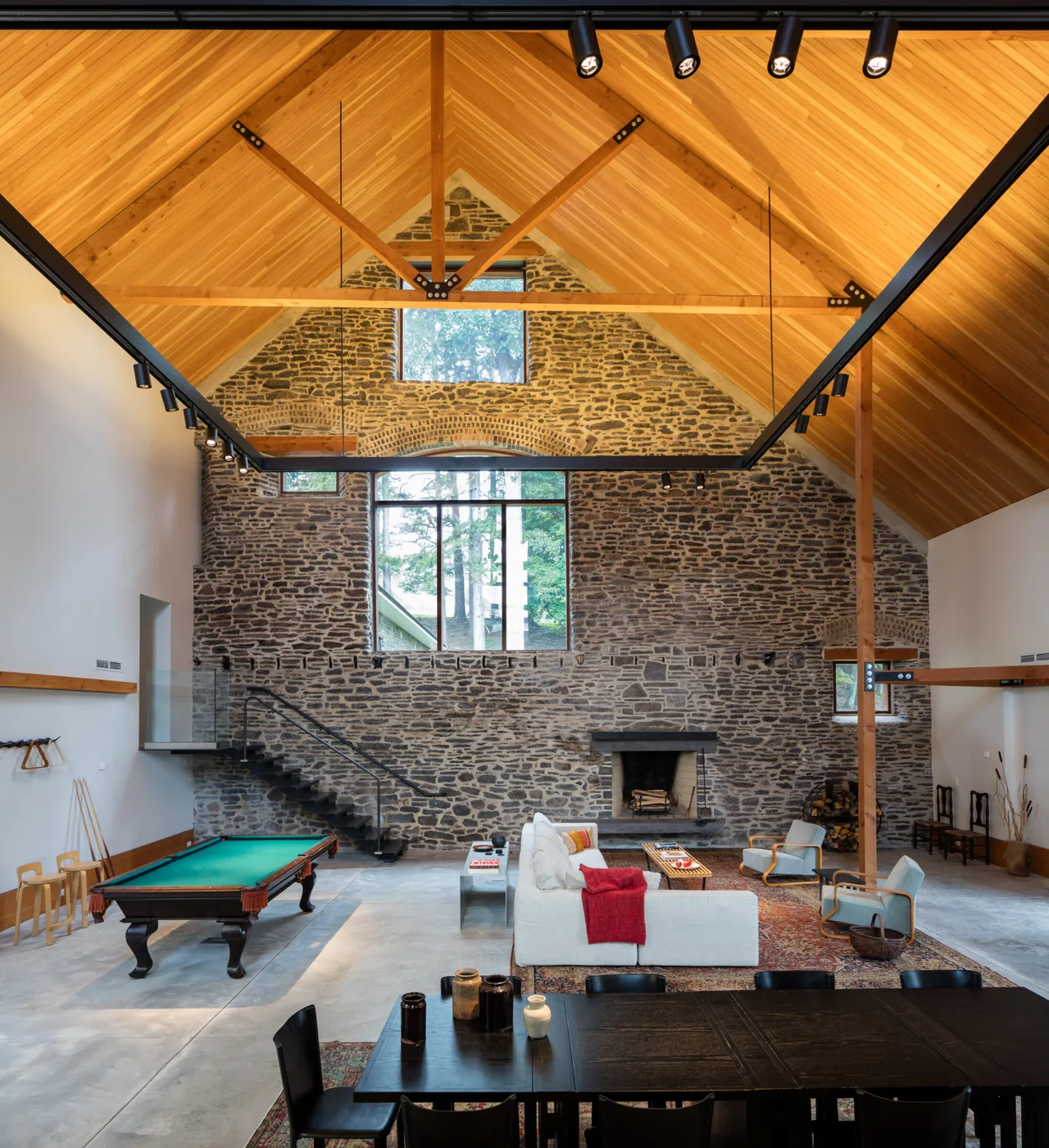 Spacious interior with vaulted wood ceiling, stone wall, staircase, fireplace, and modern furnishings in a barn-style building.