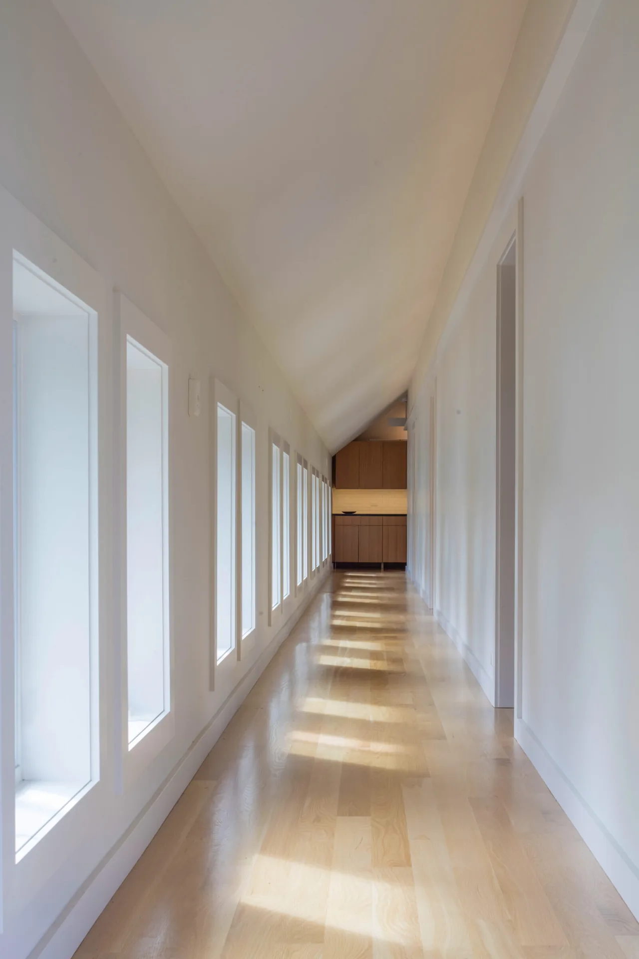 A minimalist hallway with multiple rectangular windows on one side and light wood flooring.