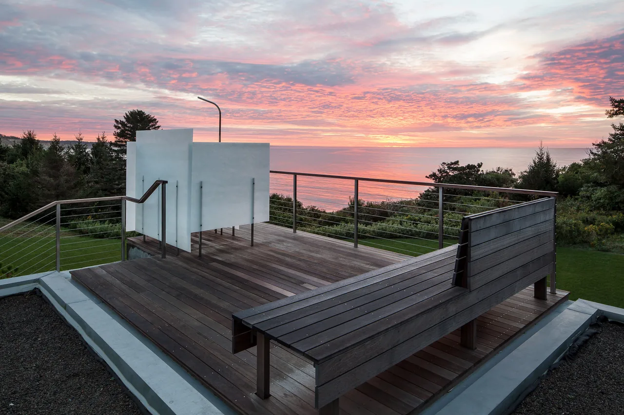 The East House features a wooden patio deck with seating and metal cable railing overlooking a coastal landscape at sunset.