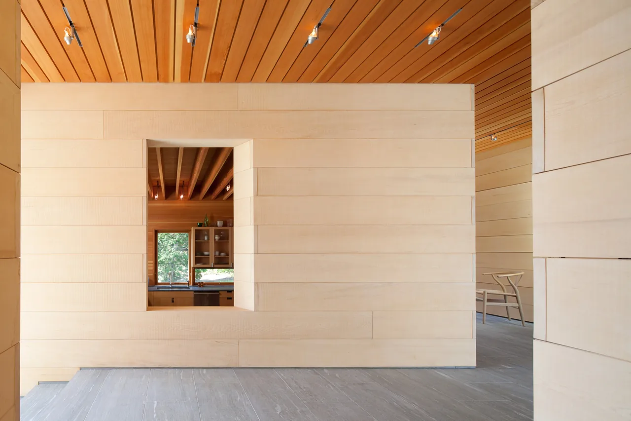 East House interior features light wood-paneled walls and ceilings with an opening overlooking the kitchen, showcasing a minimalist aesthetic.