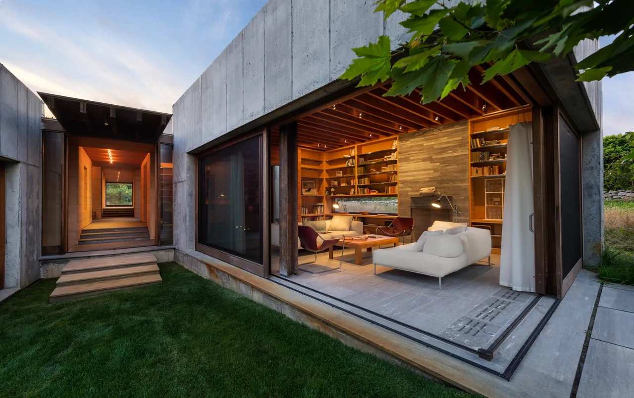 Modern house with exposed concrete walls and a living room featuring large sliding glass doors and built-in wood shelves, connected to a covered walkway.