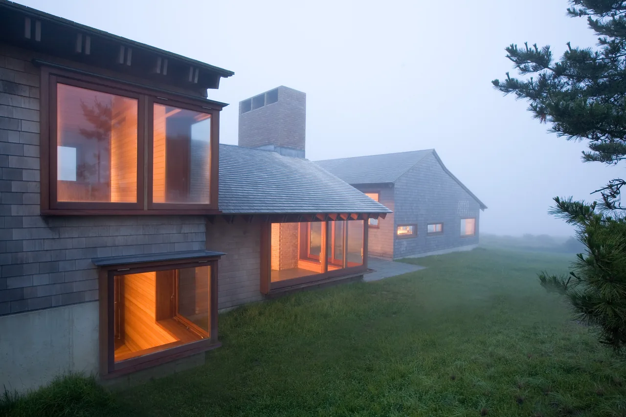 Vineyard Residence by Peter Rose + Partners features shingle siding and a brick chimney with glowing interior wood elements.