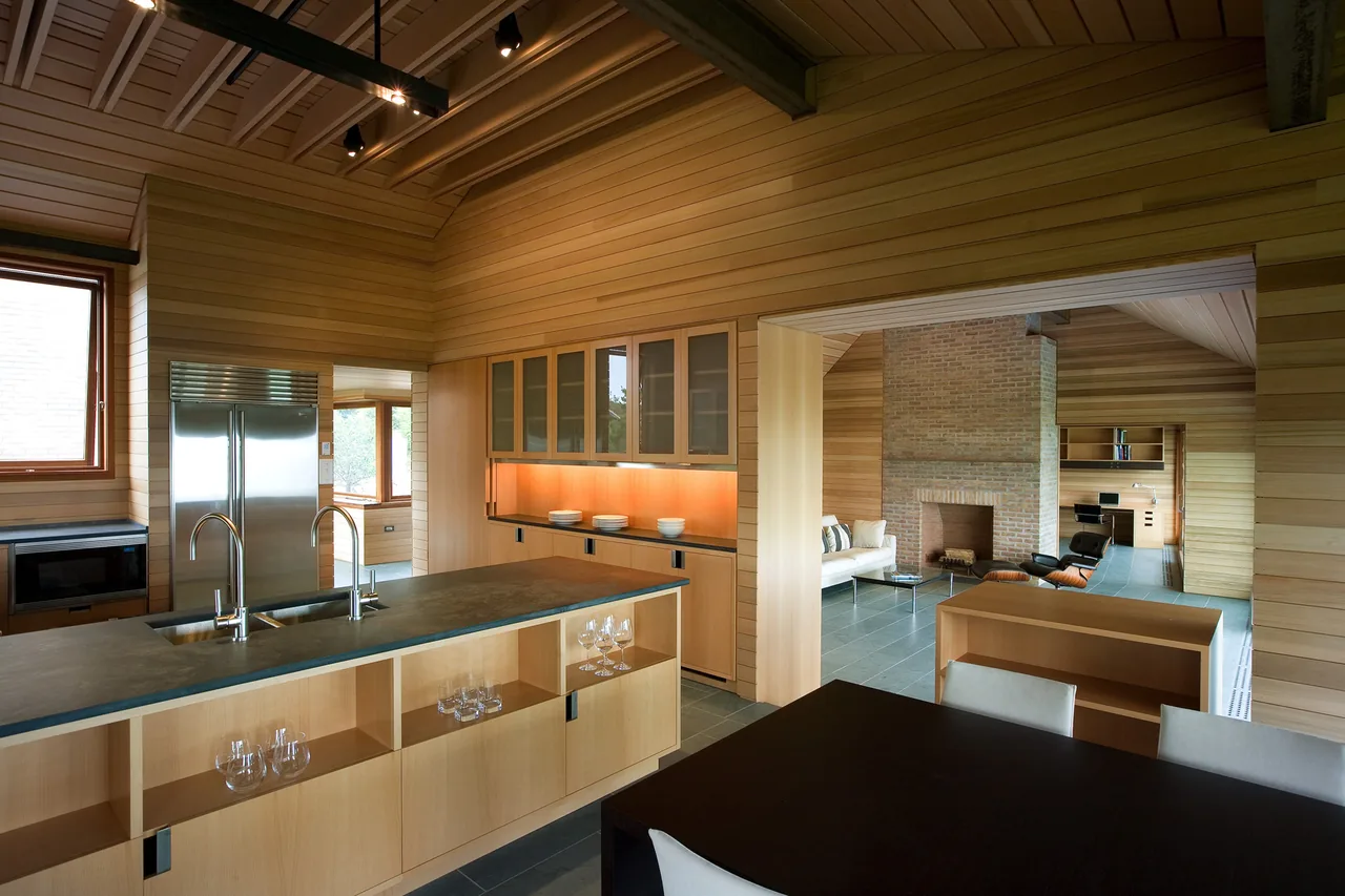 Open kitchen and living room area with warm wood paneling, stone tile flooring, and a brick fireplace in the Vineyard Residence.