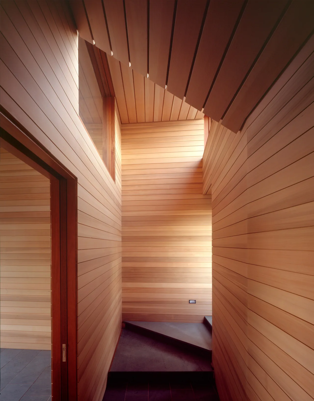 Interior view of a wood-paneled stairwell with triangular concrete steps and a high window.