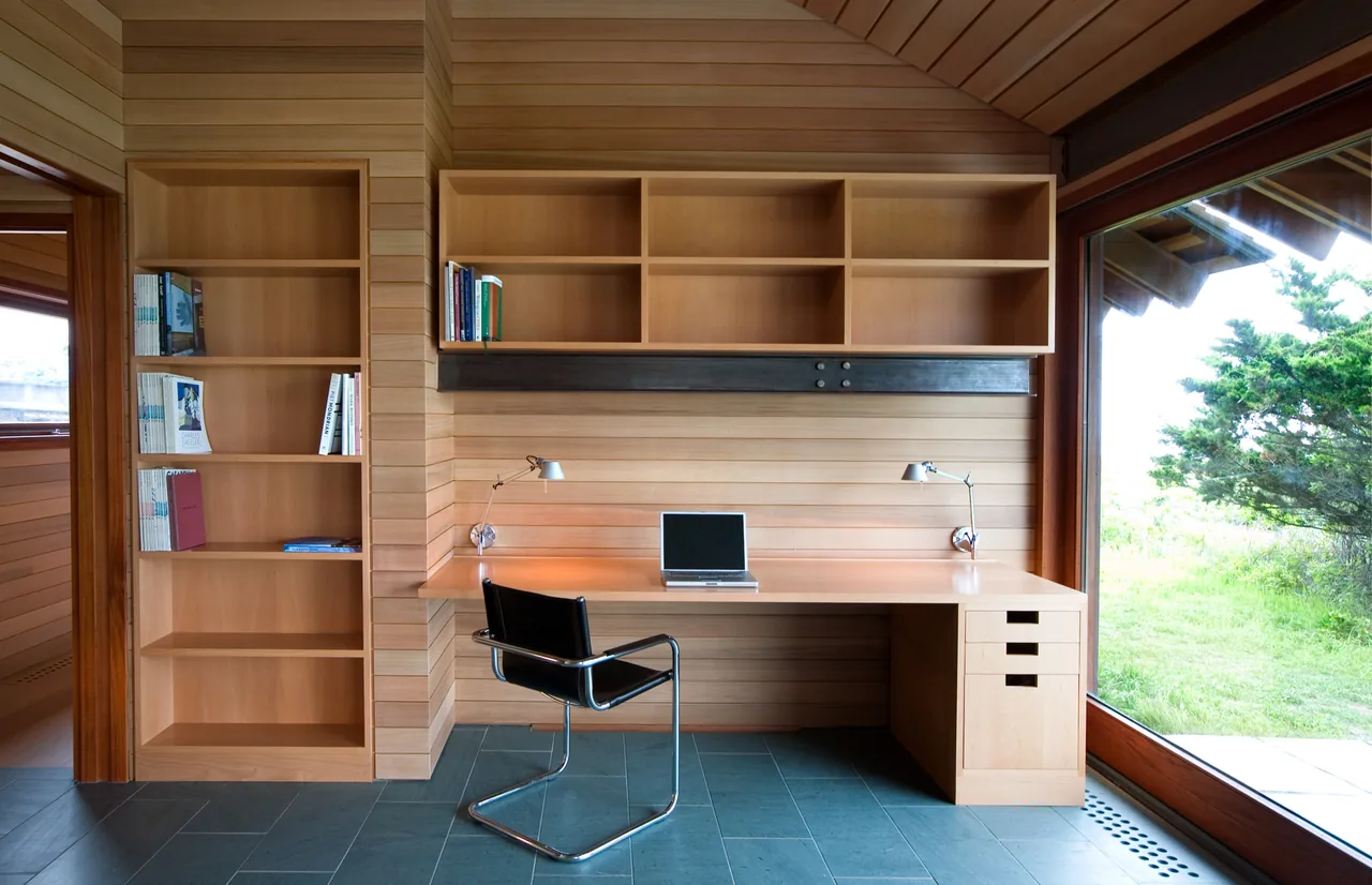 Home office with wood-paneled walls and ceiling, built-in shelving, slate tile floor, and large wood window frames overlooking the landscape.