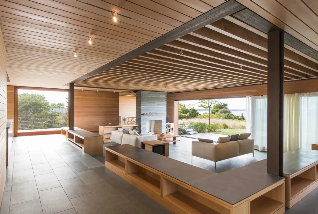 The Island Residence living room by Peter Rose + Partners features wood slat ceilings, a stone fireplace, and large openings to the outdoors.