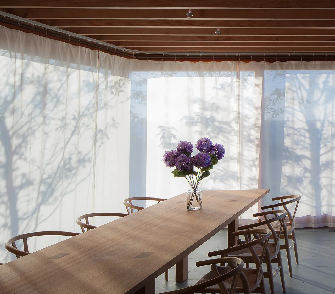 Dining area with a long wooden table, six chairs, and sheer curtains creating soft natural light effects.