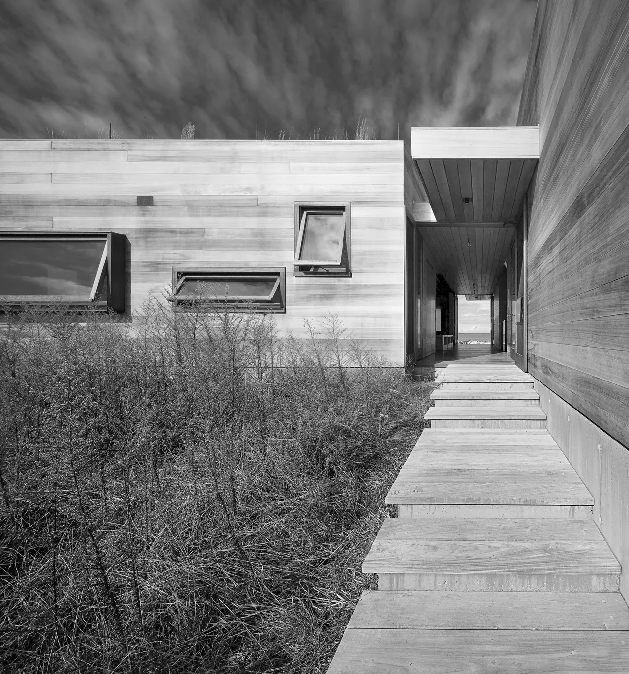 Modern residence featuring flat roof, raised foundation, wooden siding, and large rectangular hinged windows with a steel railing visible.